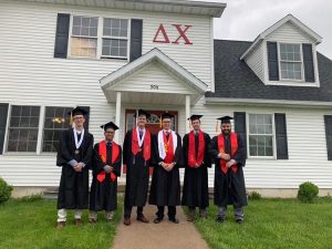 Six graduates in caps and gowns standing in front of a fraternity house.