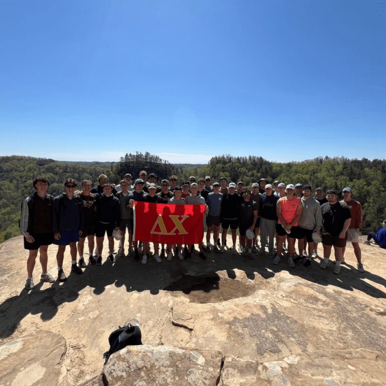 A large group of people posing outdoors on a rock with a red flag under a clear blue sky.