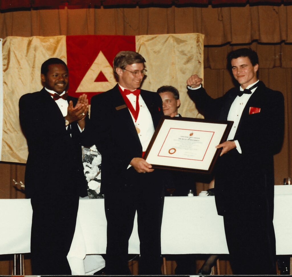 Three men in tuxedos holding a framed certificate on stage.