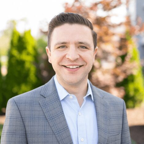 Smiling man in suit outdoors with trees.