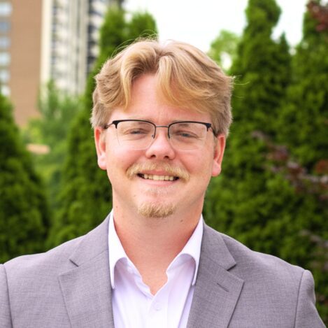 Smiling man in gray suit with glasses outdoors.